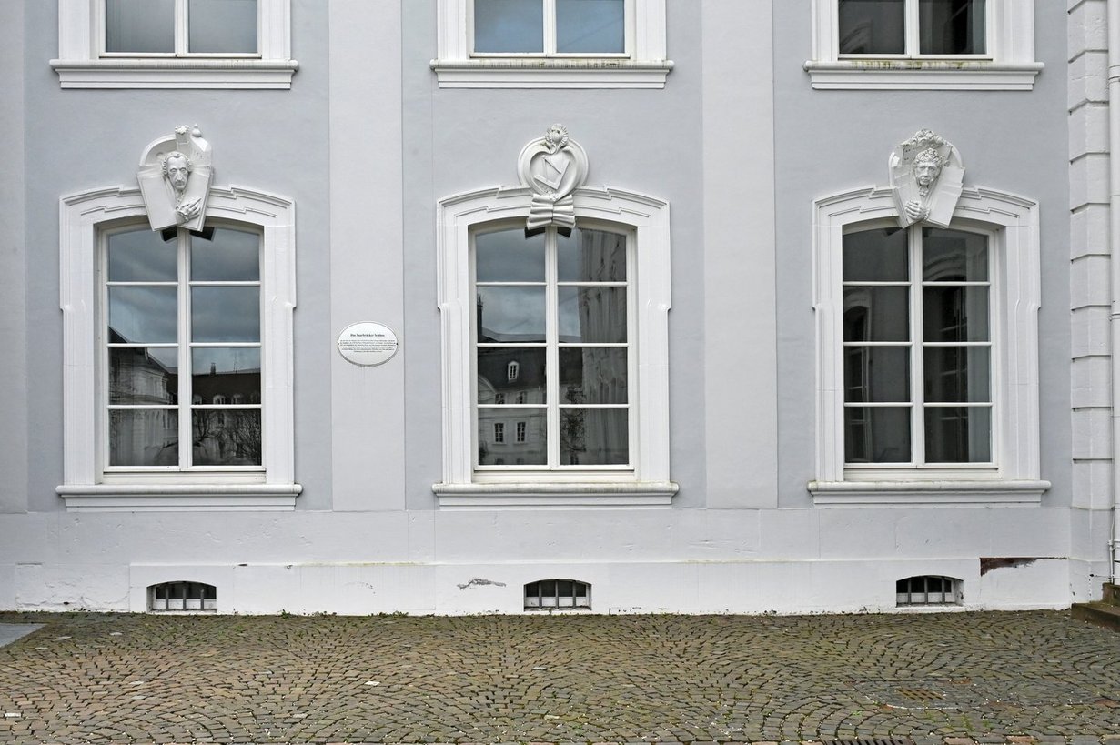 Blick auf die weißen Fensterrahmen dreier Fenster im Erdgeschoss des Saarbrücker Schlosses. An zwei Fensterstürzen in Weiß die plastischen Porträts der Architekten Friedrich Joachim Stengel und Johan Adam Knipper dem Jüngeren. Auf dem mittleren Relief die Insignien Winkel und Zirkel.